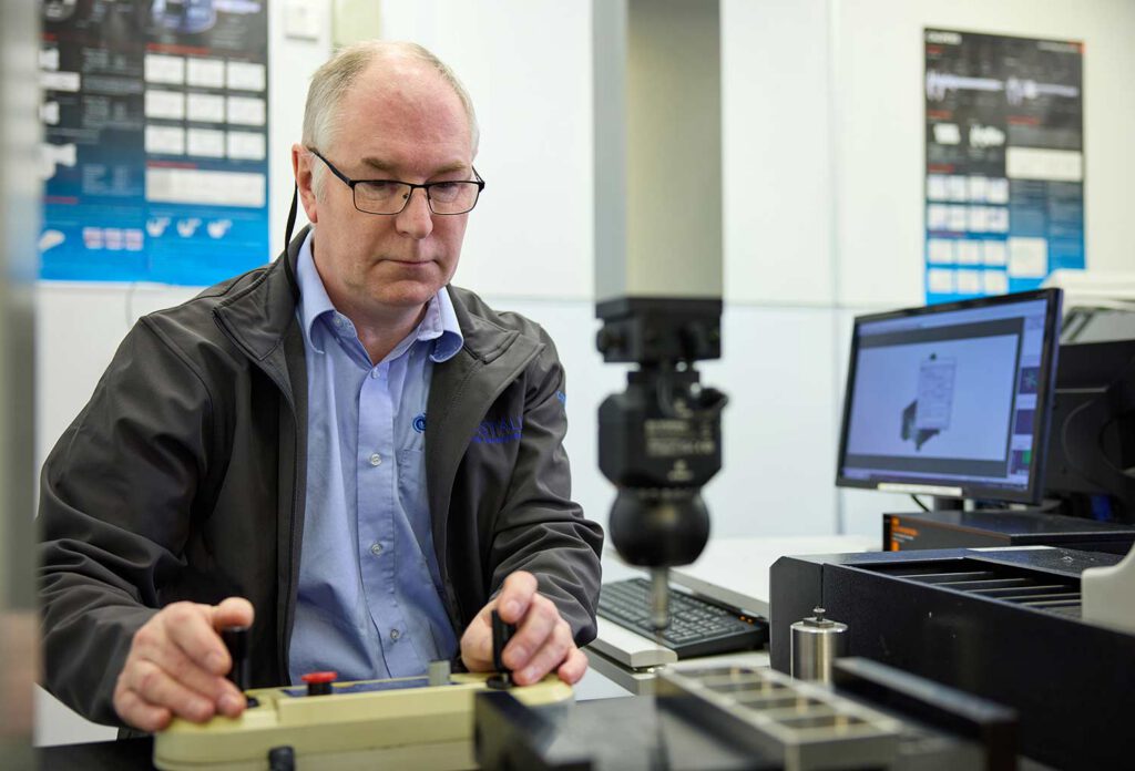Mark and the CMM (coordinate measuring machine) - A man with grey hair and glasses has his hands on the joystick controls of a device, a machine part can be seen defocused to the front of the image, a computer screen is seen to the middle right