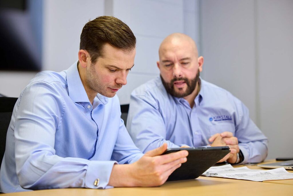 Two men, wearing blue shirts, look at a tablet held by the man to the left.