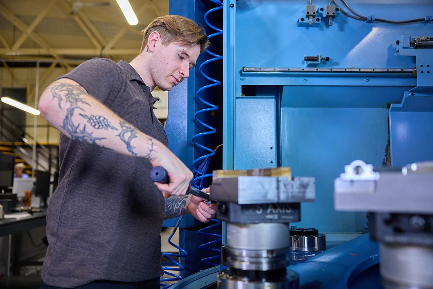 A young man with mid brown/blond hair works at a blue machine, he has tattoos visible on his right arm.